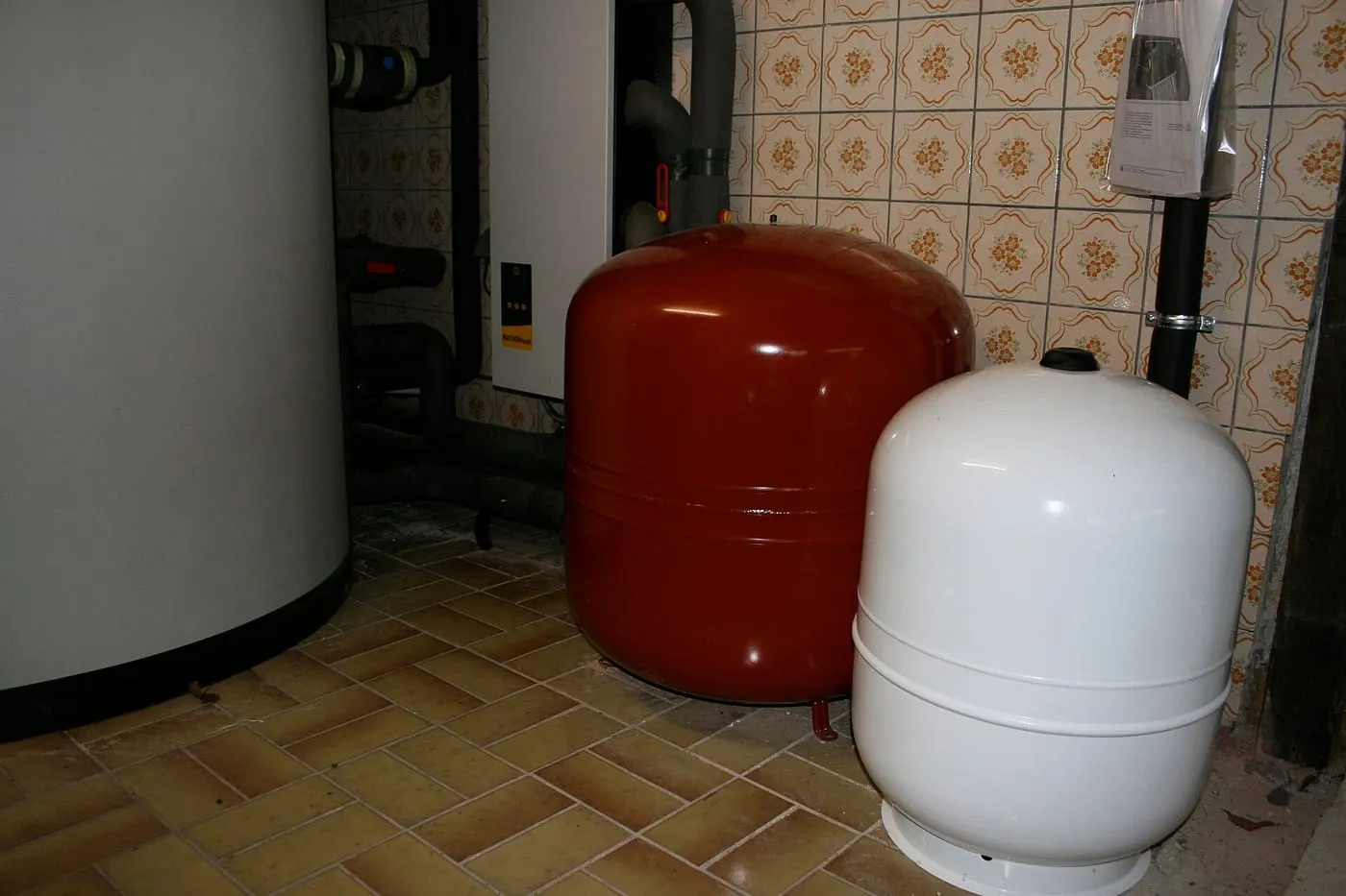 Red and white expansion vessels standing on a tiled floor beside a hot water tank