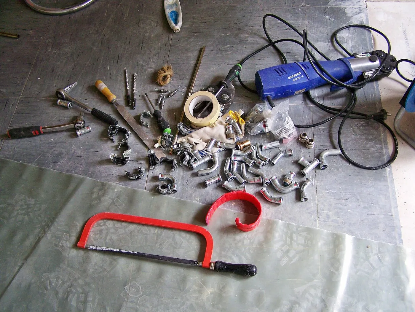 Plumbing tools and fittings laid out on a dust sheet, with a hacksaw and pipe bender