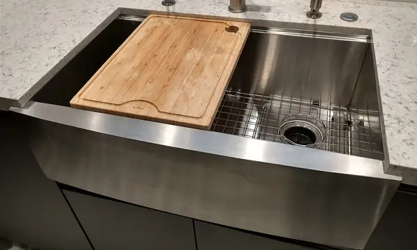 A stainless steel farmhouse kitchen sink with a wooden chopping board and mixer tap