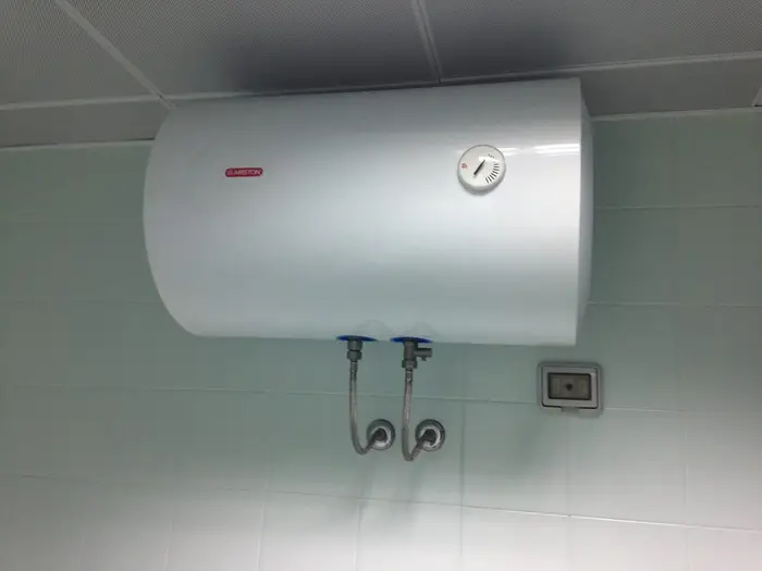 A horizontal electric water heater mounted high on a tiled wall with its flexible connections