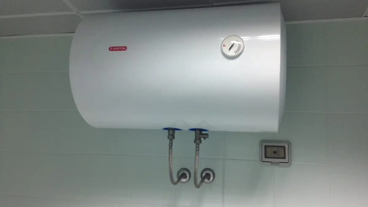 A horizontal electric water heater mounted high on a tiled wall with its flexible connections